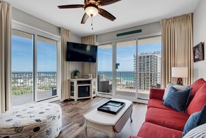 Un salón amplio con un sofá rojo, una mesa de centro blanca y un televisor de pantalla plana. La habitación tiene ventanas grandes con vistas a la ciudad y un balcón.