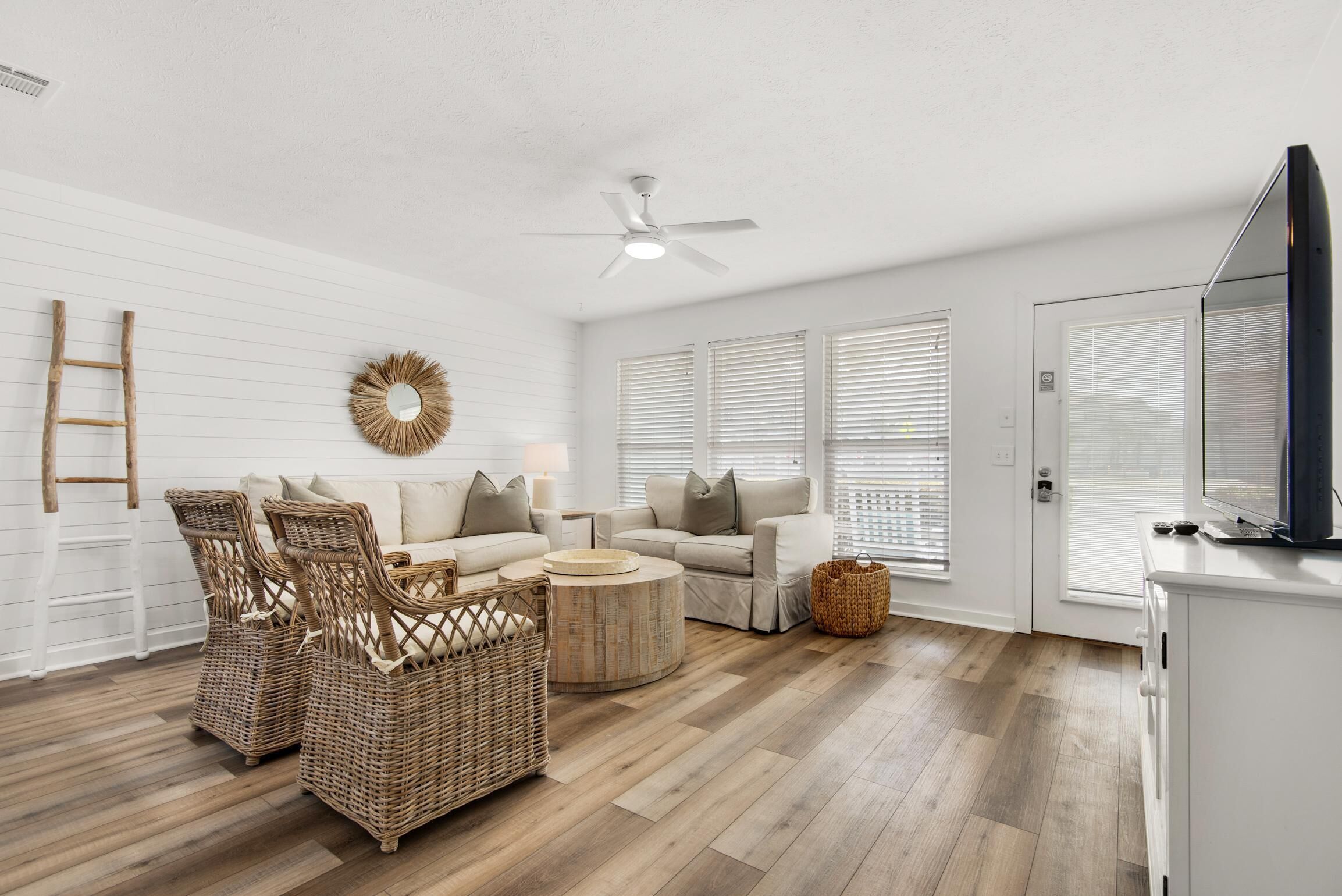 A modern living room with a white wall, a ceiling fan, wicker furniture, a round coffee table, and a television on a cabinet.