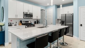A modern kitchen with white cabinets, stainless steel appliances, a central island with bar stools, and pendant lighting. - Townhome with Screened Patio, Games Area & Close to Pool (Kissimmee)