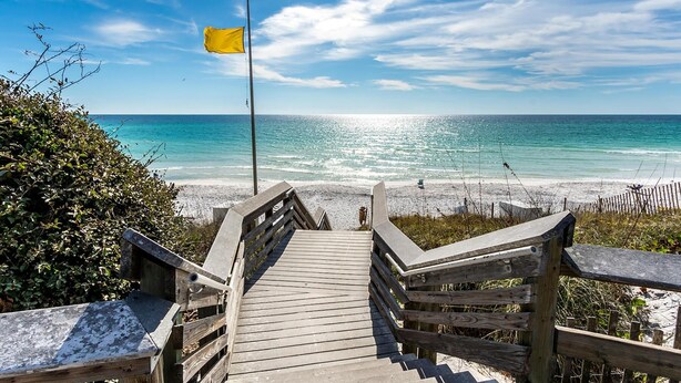 Un escalier en bois mène à une plage où flotte un drapeau sur un mât.