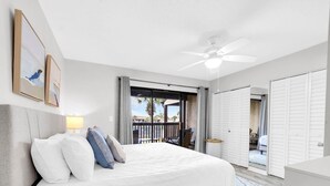 A bedroom with a bed, a nightstand, a ceiling fan, a window with a view, and a door leading to another room. - Portside R4 (Panama City Beach)