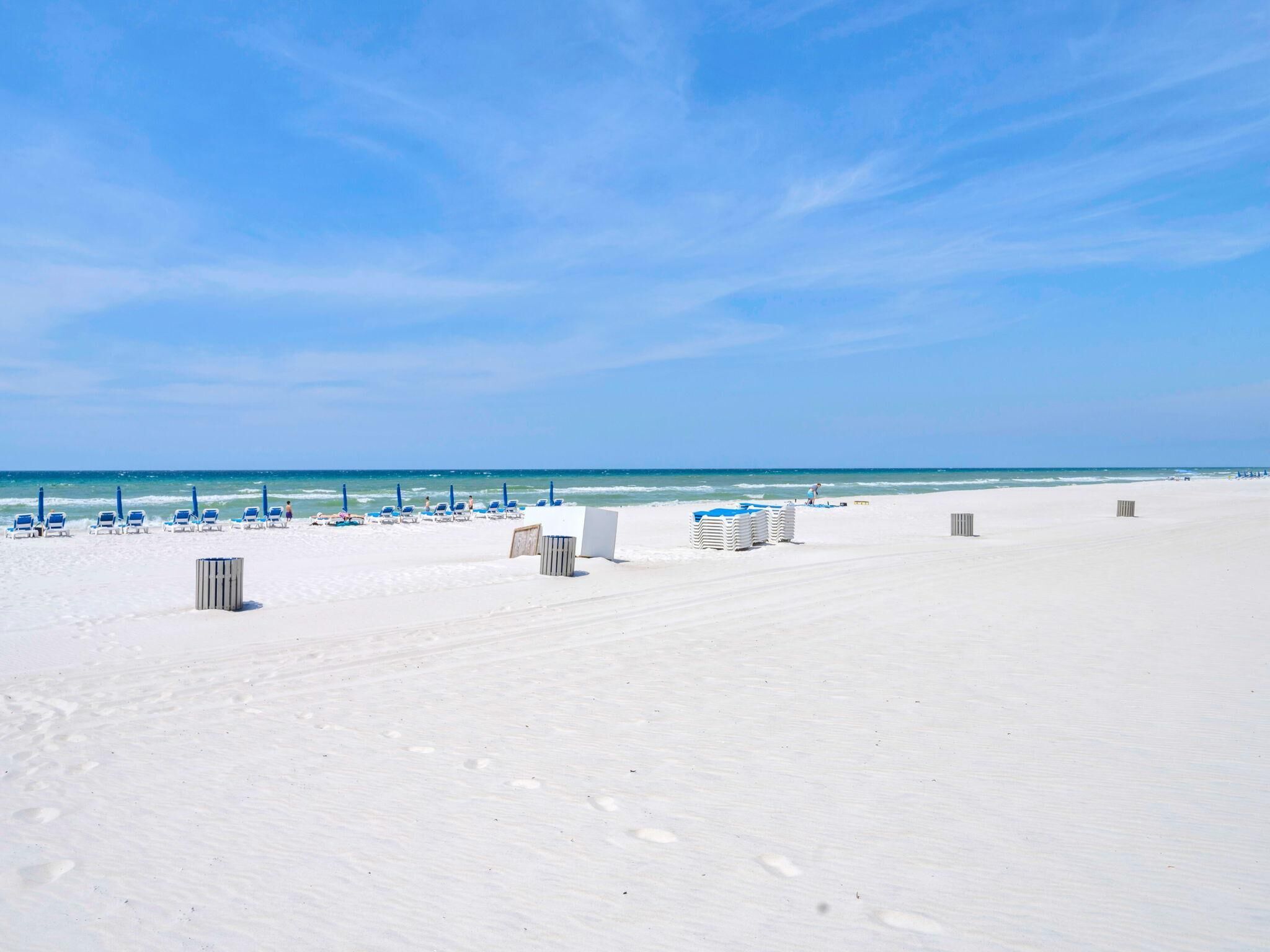 Una spiaggia con sabbia bianca, cielo azzurro limpido e oceano. Si vedono anche poltrone da spiaggia blu e un edificio bianco.