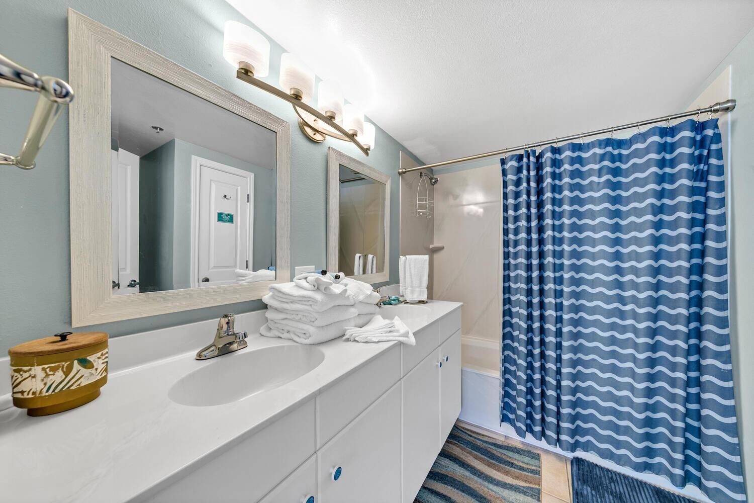 Un baño con tocador blanco, un espejo grande, un perchero y una cortina de ducha con diseño azul ondulado.