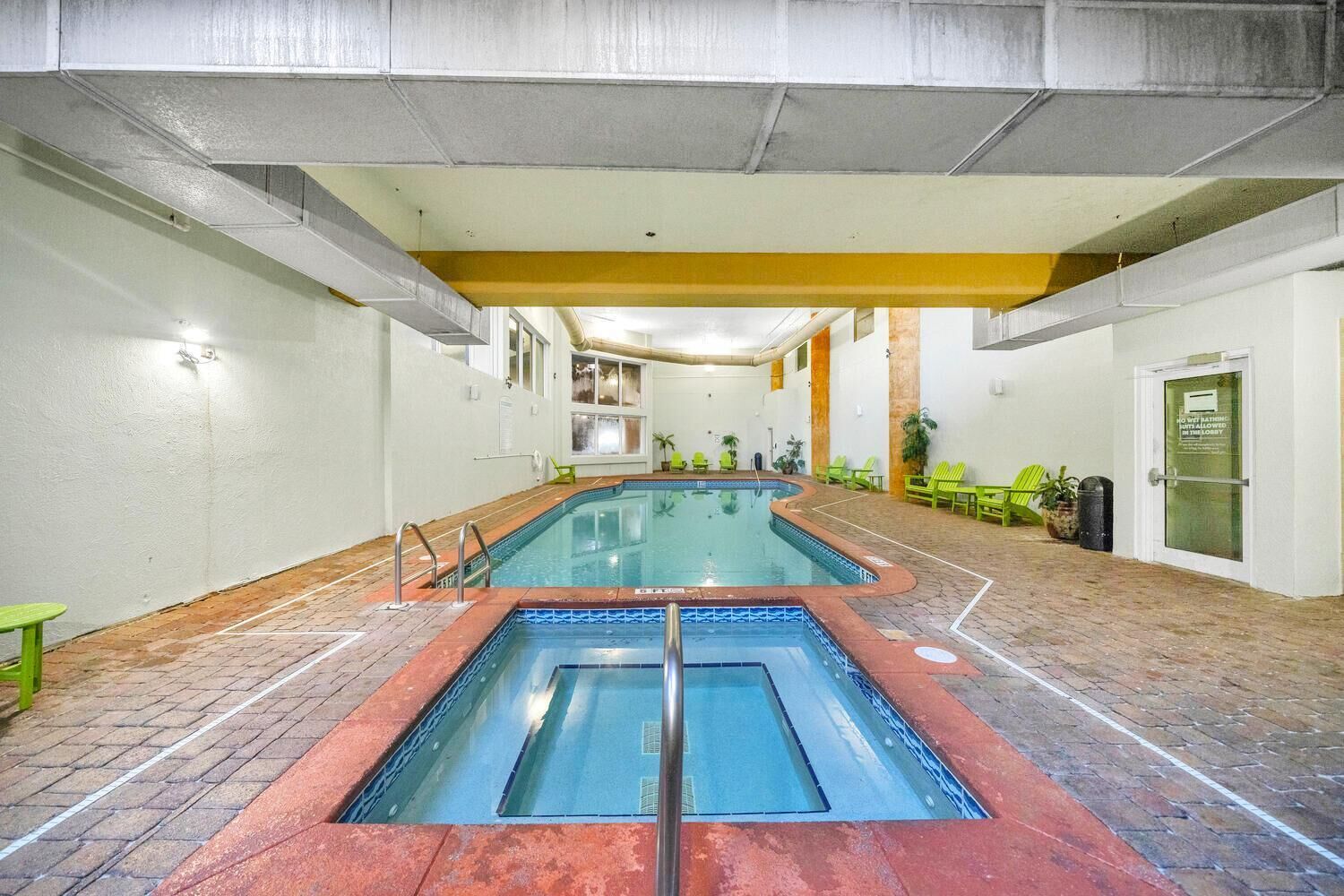 Une piscine intérieure avec deux couloirs, entourée d’un sol en briques et de chaises vertes.
