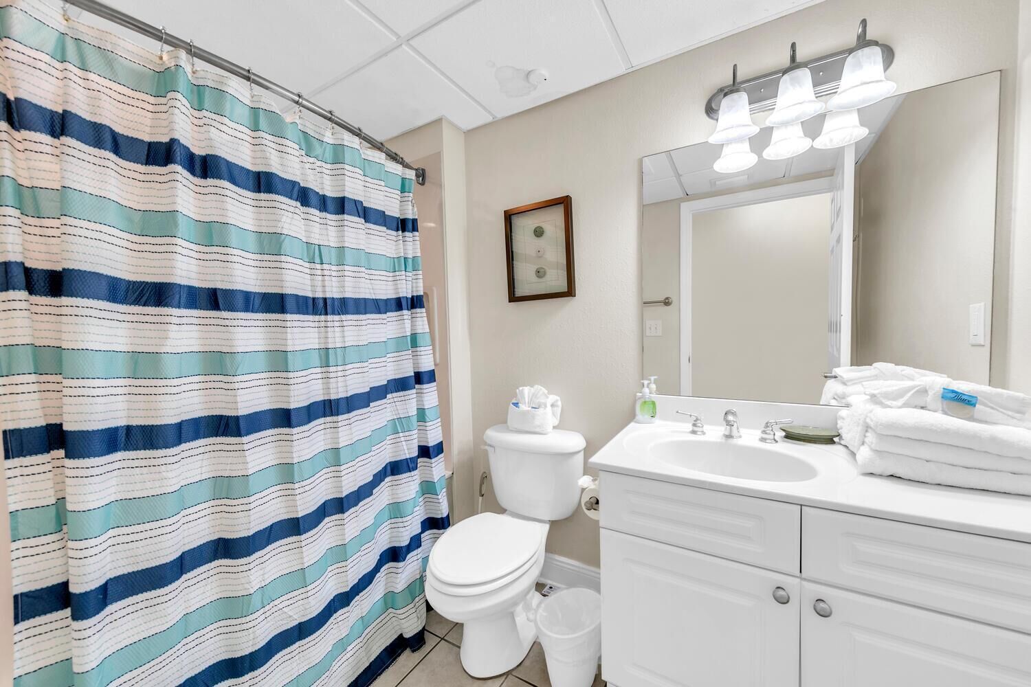 A bathroom with a striped shower curtain, a toilet, a sink with a mirror, and a trash bin.