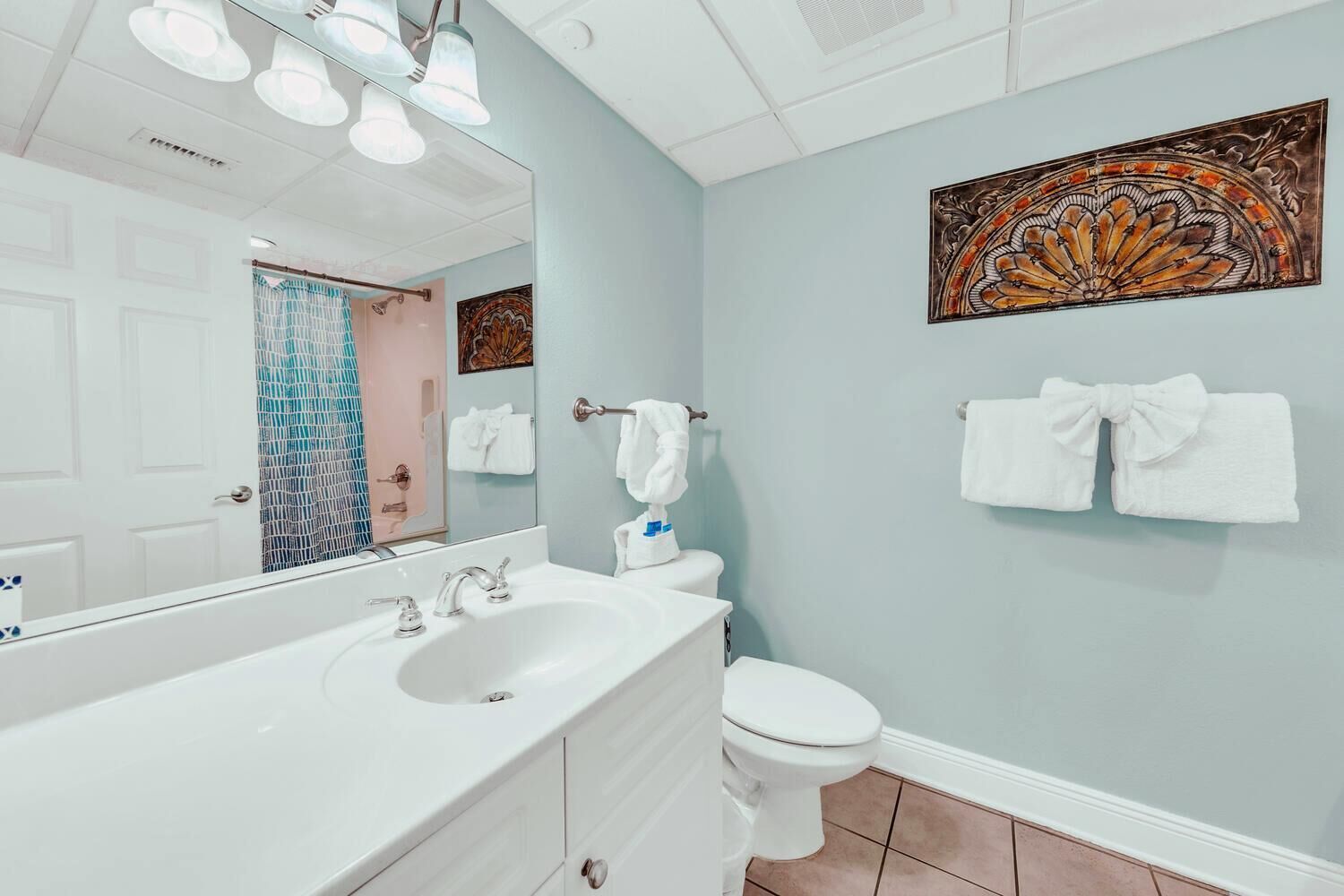 A bathroom with a white sink, two faucets, a mirror, a toilet, and a shower curtain. There are towels hanging on a rack and a decorative wall piece above the toilet.