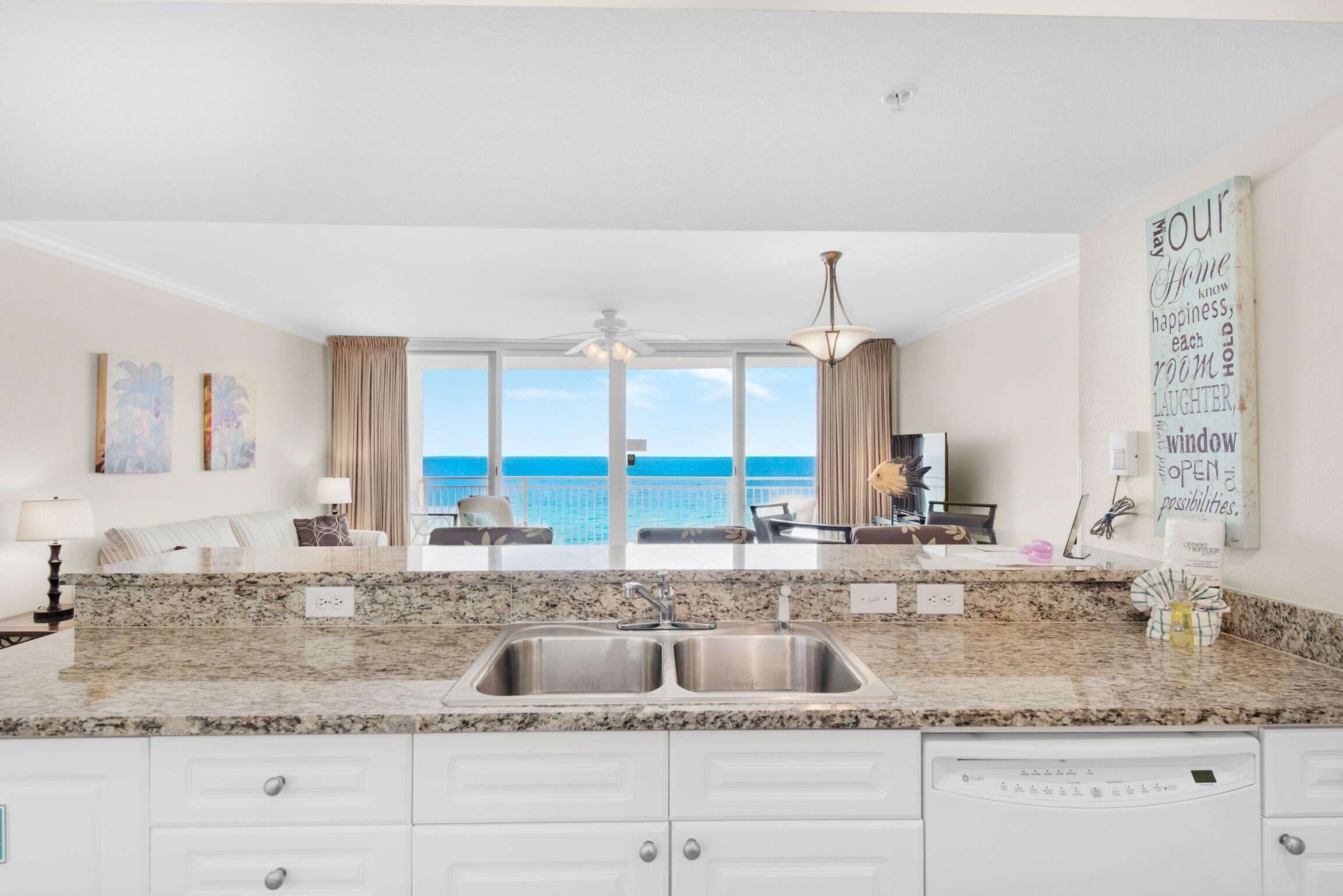 A kitchen with a granite countertop, double sink, and white cabinetry. There is a view of the ocean through large windows.