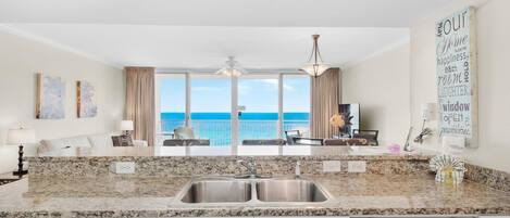 A kitchen with a granite countertop, double sink, and white cabinetry. There is a view of the ocean through large windows.