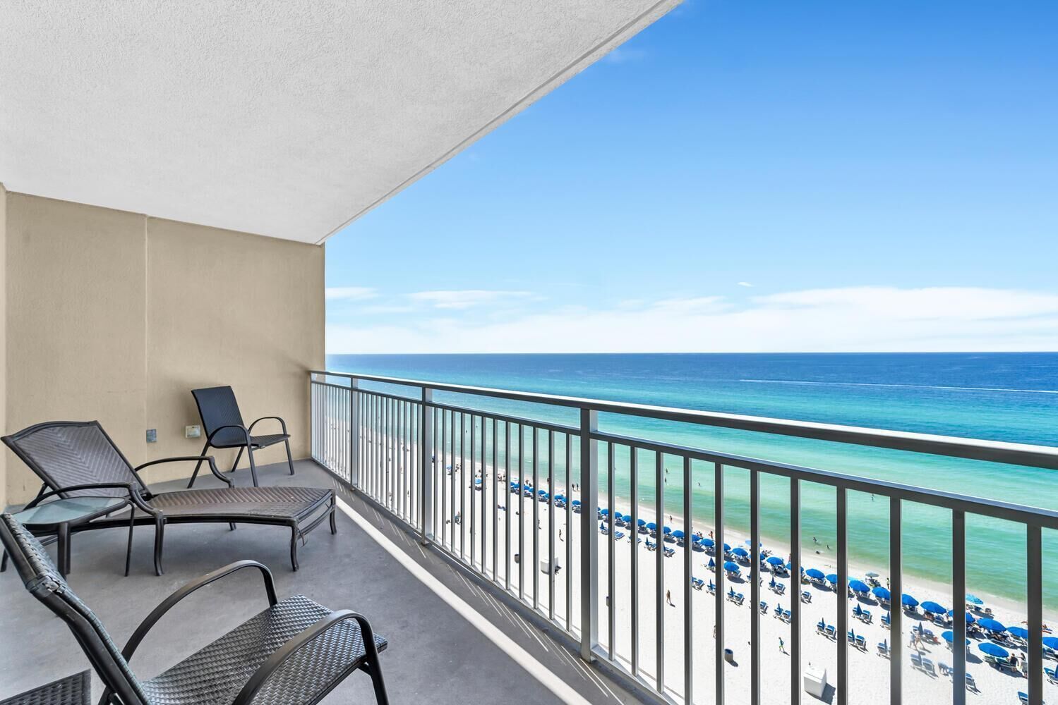 Un balcon avec deux chaises et une table, donnant sur une plage parsemée de parasols et où se trouvent des gens.