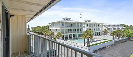 A balcony with a view of a multi-story building, a swimming pool, and parked cars.