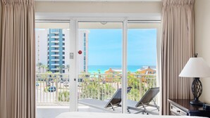 A hotel room with a bed, a nightstand with a lamp, a balcony with a view of the ocean and buildings, and a sliding door. - Sterling Shores 402 (Destin)