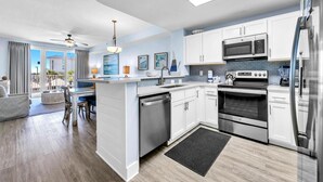 A modern kitchen with white cabinets, stainless steel appliances, and a dining area with a view. - Calypso 3-202 - The Sea Suite (Panama City Beach)