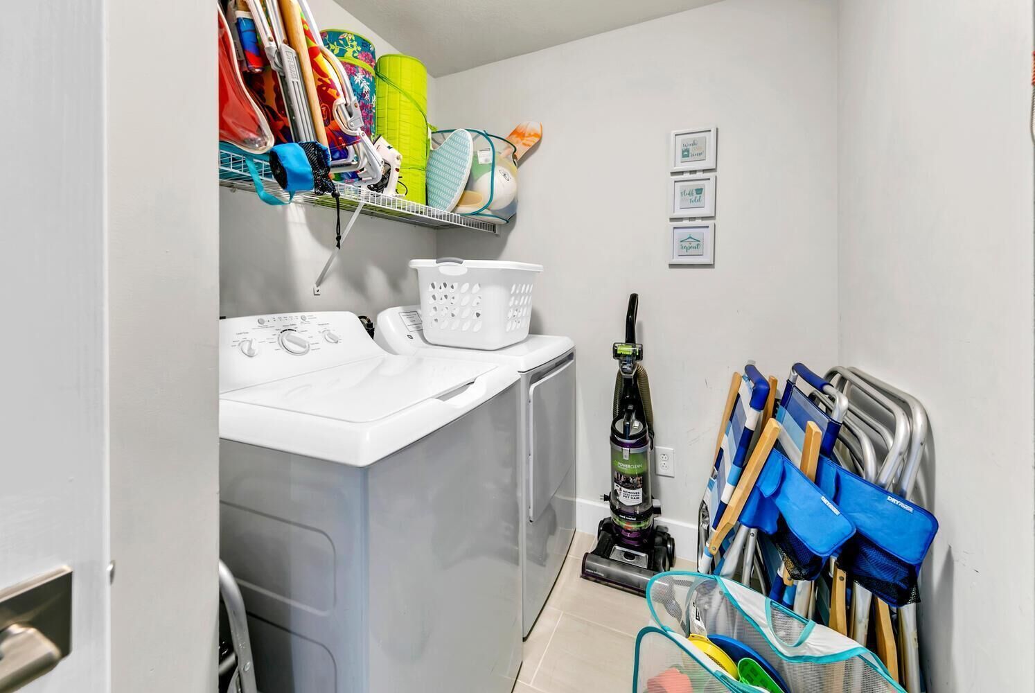 A laundry room with a washing machine, dryer, and a vacuum cleaner. There are baskets and cleaning supplies on shelves.