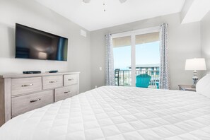 A modern hotel room with a large bed, a flat-screen TV mounted on the wall, a dresser with drawers, a balcony with a view, and a nightstand with a lamp. - Calypso 3-406 - Paradise by the Pier (Panama City Beach)