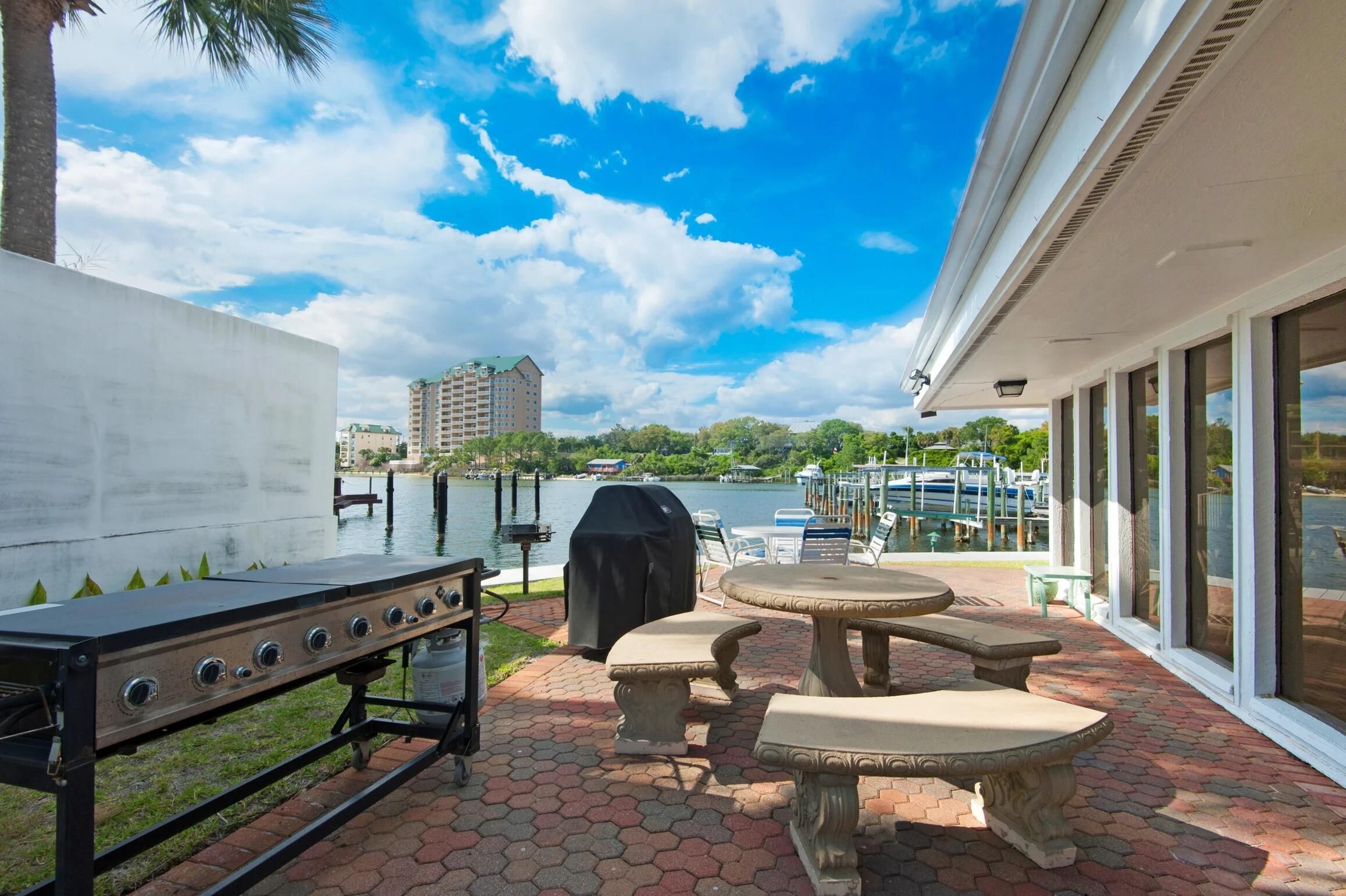 An outdoor patio with a grill, tables, and chairs overlooking a lake with boats and buildings in the background.