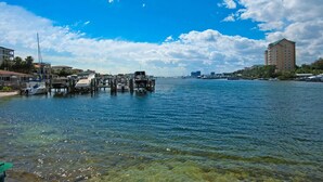 Un puerto deportivo frente al mar con barcos amarrados a muelles de madera, un cielo azul despejado y edificios al fondo.