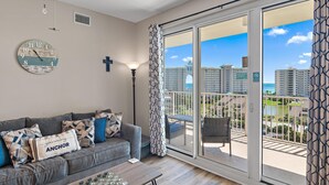 A living room with a grey sofa, patterned cushions, a round clock, and a view of a balcony with outdoor furniture. - Ariel Dunes II 703 (Miramar Beach)