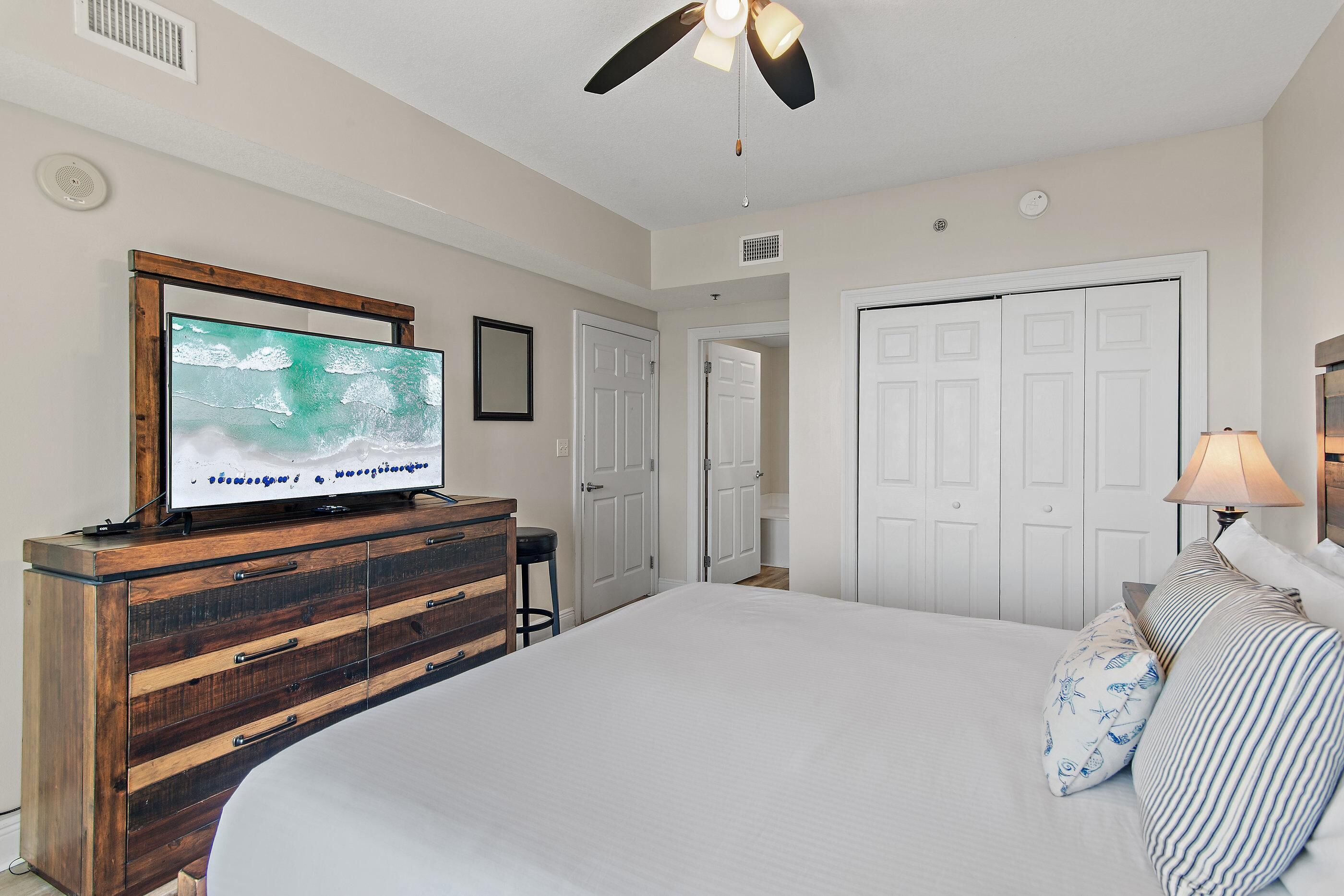 A bedroom with a bed, a television on a wooden dresser, a ceiling fan, and white doors.
