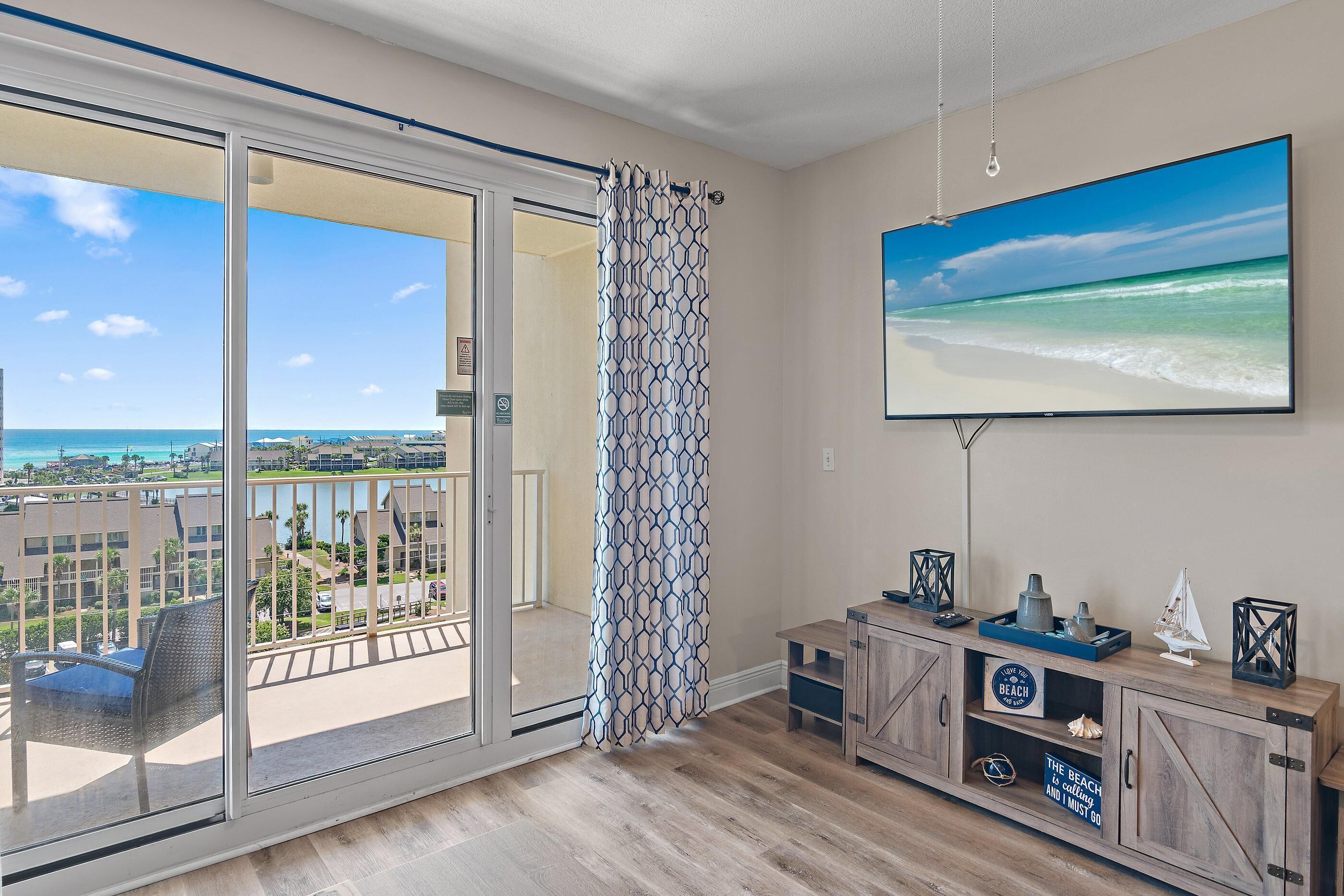 A balcony with a view of the beach, a sliding glass door, a wooden cabinet with decorative items, and a large TV screen displaying a beach scene.
