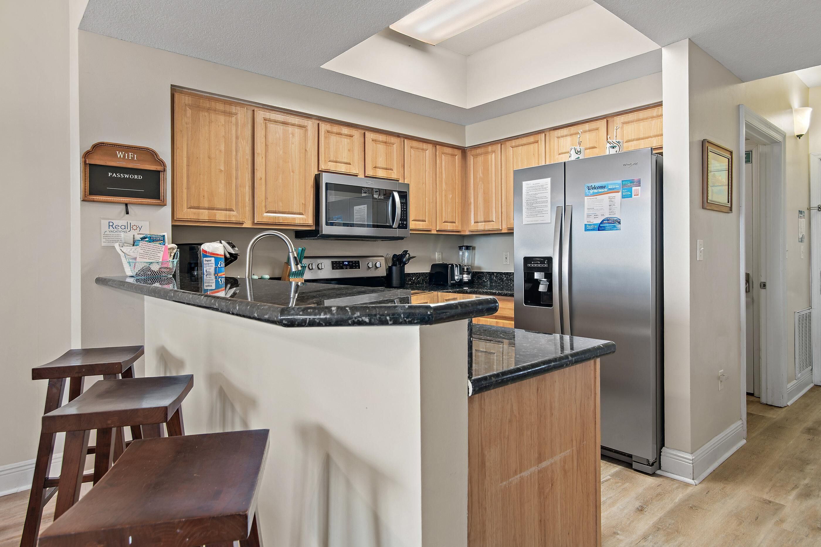 A modern kitchen with wooden cabinets, a built-in microwave, a stainless steel refrigerator, and a small island with stools.
