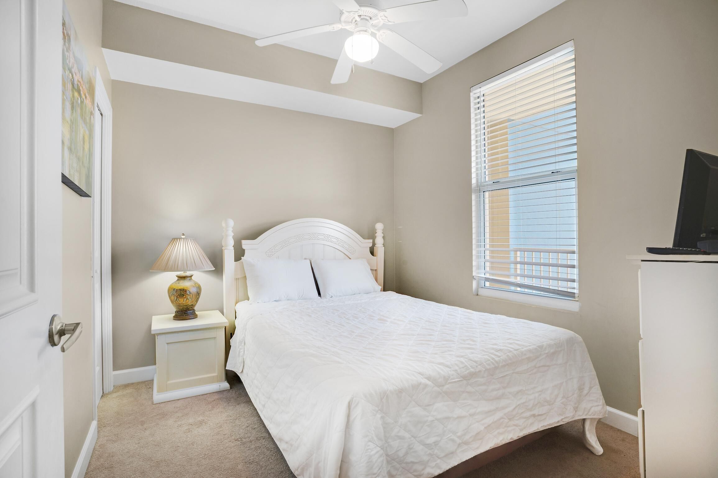 A bedroom with a bed, a nightstand with a lamp, a ceiling fan, and a window with blinds.