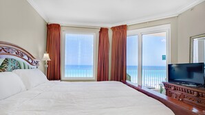 Ein großes Bett mit Holz Kopfteil, ein Fernseher auf einem Holzgestell und Meerblick durch große Fenster.