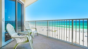Ein Balkon mit zwei Adirondack-Stühlen, einer Glastür und Blick auf einen Strand mit blauem Himmel.