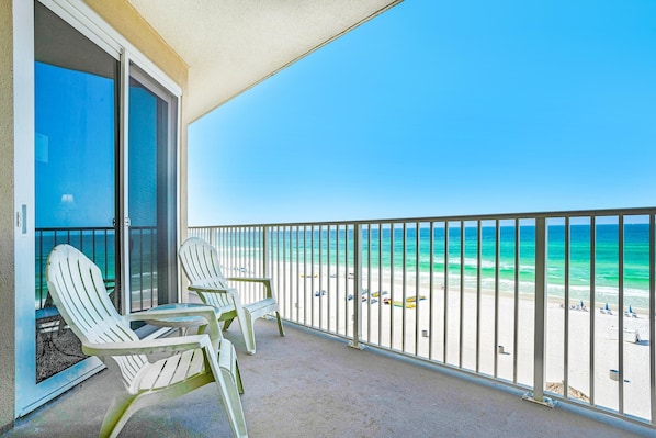 A balcony with two Adirondack chairs, a glass door, and a view of a beach with a clear blue sky.