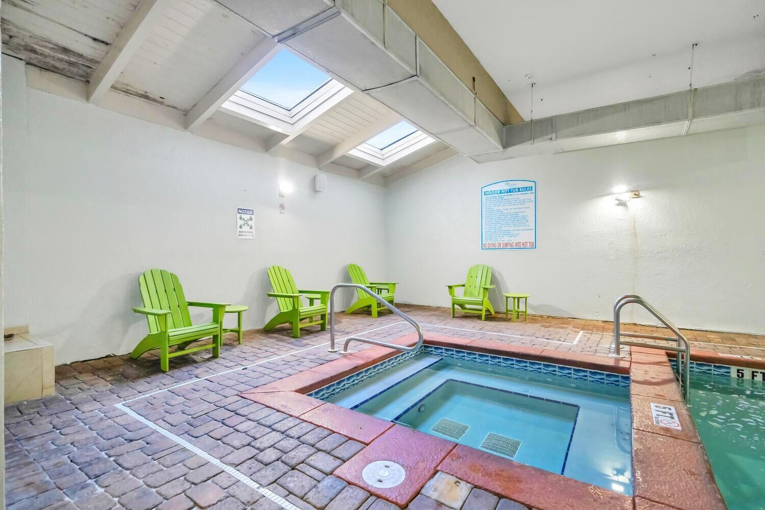 An indoor swimming pool with a skylight ceiling, green Adirondack chairs, and a safety sign.