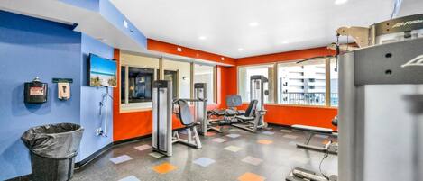 Un gimnasio con equipo de ejercicio, un bote de basura y una ventana con vistas a edificios.