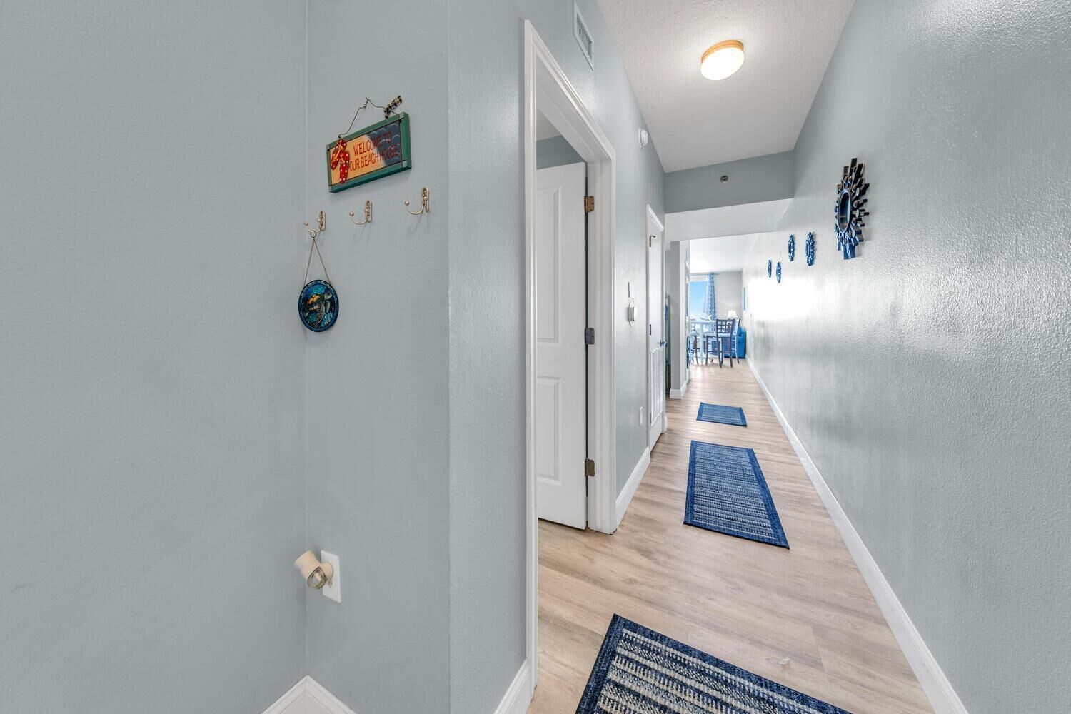 Un couloir aux murs bleu clair, doté d’un plancher en bois et de tapis bleus. Des portes-fenêtres donnent accès à d’autres pièces, et un panneau est fixé au mur.