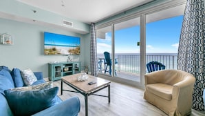 A living room with a large window, a painting of a beach, a teal cabinet, and a beige armchair. - Shores of Panama 830 (Panama City Beach)