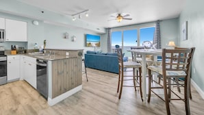 A modern kitchen with a central island, a dining area with wooden chairs, and a view of the ocean through large windows. - Shores of Panama 830 (Panama City Beach)