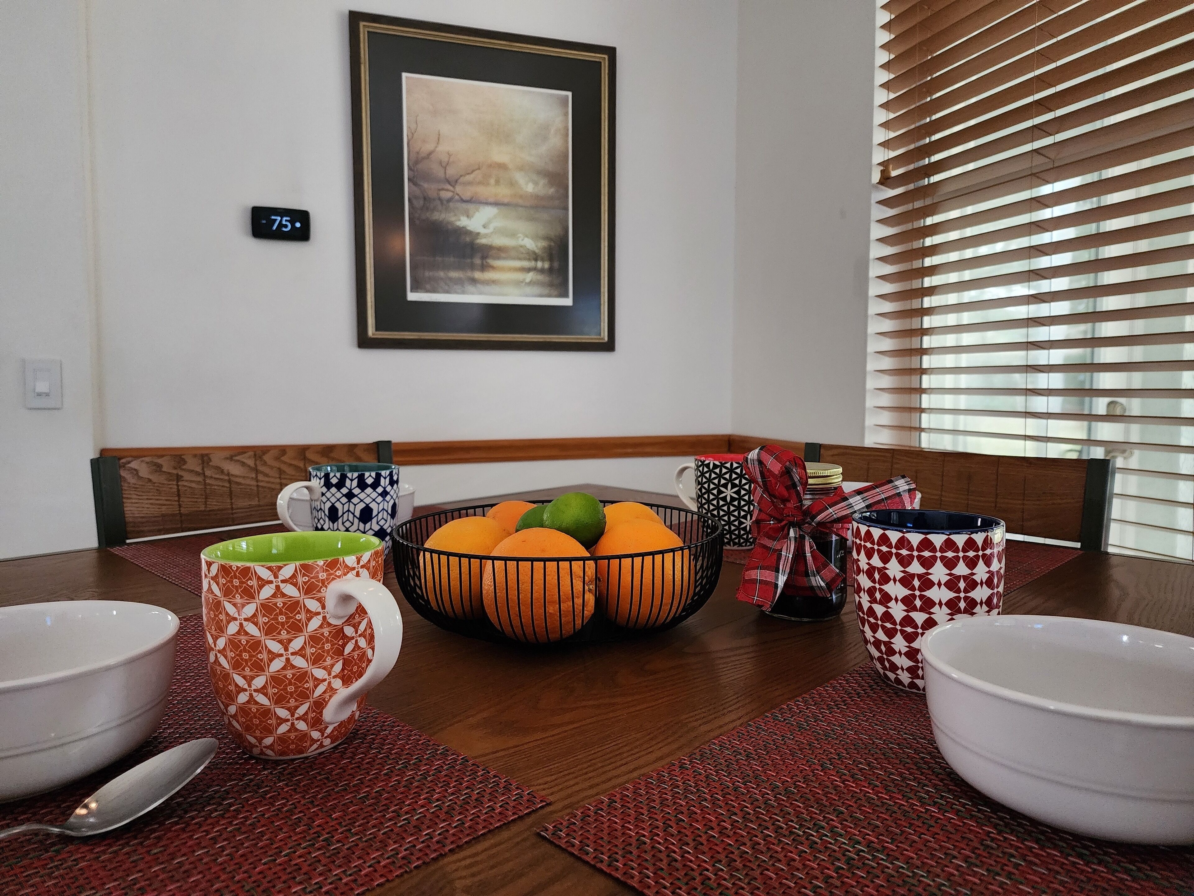 A dining table with bowls, a fruit bowl, and patterned mugs. There is a framed picture on the wall and a thermostat.