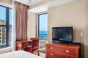 A hotel room with a large window, a bed, a desk with a television, and a view of the ocean.