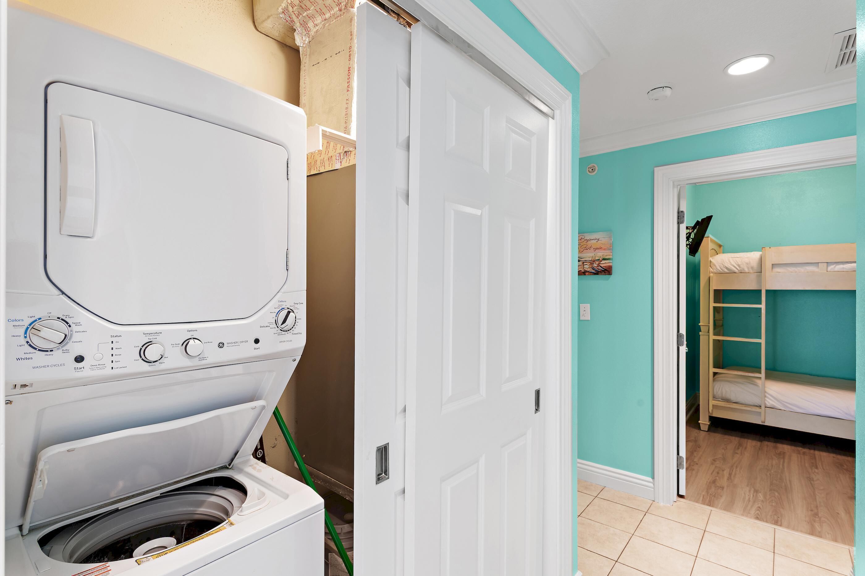 Une buanderie équipée d’un lave-linge, avec une porte donnant sur une pièce où se trouve un lit superposé et des murs de couleur turquoise.