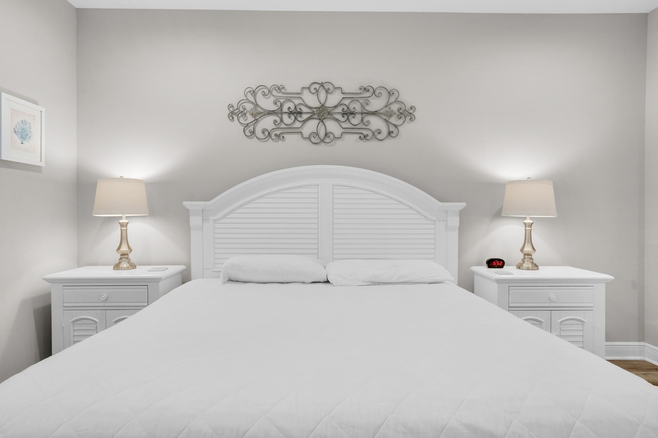 A bedroom with a white bed, two bedside tables with lamps, a decorative wall piece, and a small clock on one table.