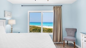 A bedroom with a large bed, a chair, a nightstand, a lamp, and a view of the beach.