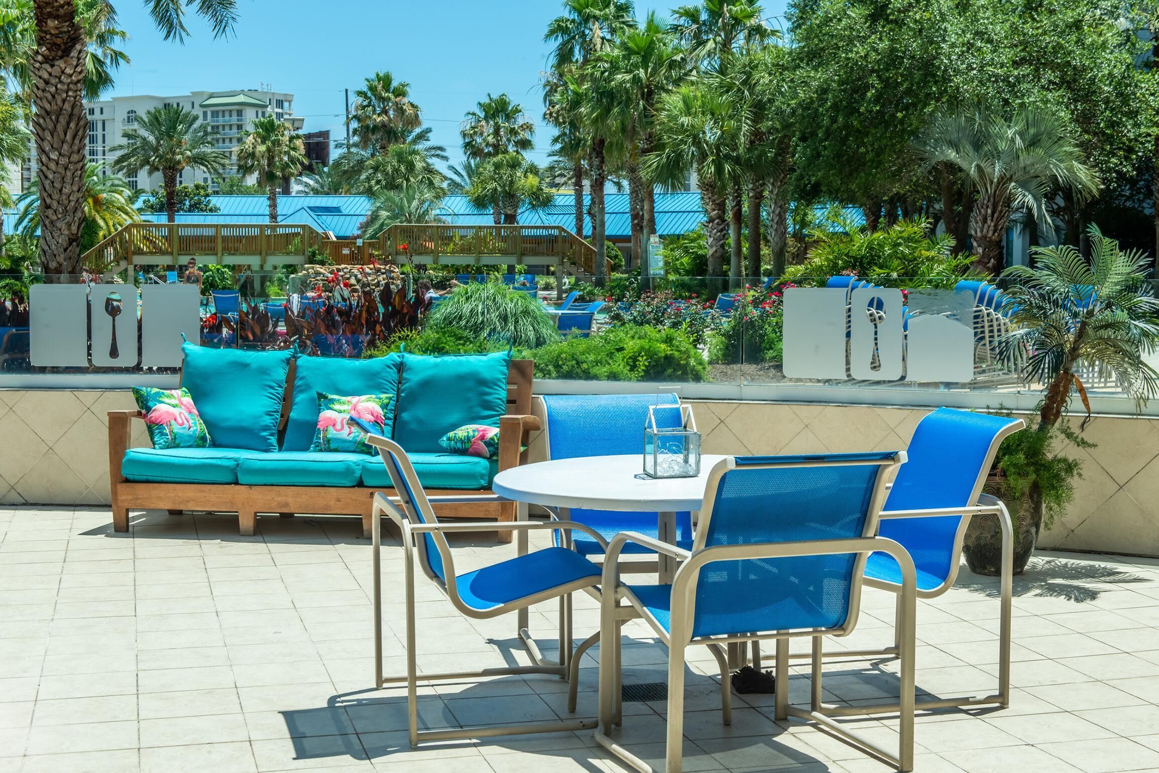 An outdoor seating area with a turquoise sofa and blue chairs, surrounded by palm trees and a view of a resort.