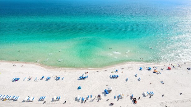 Une vue aérienne d’une plage avec des rangées de transats et de parasols, une mer bleue et limpide, et une plage de sable.