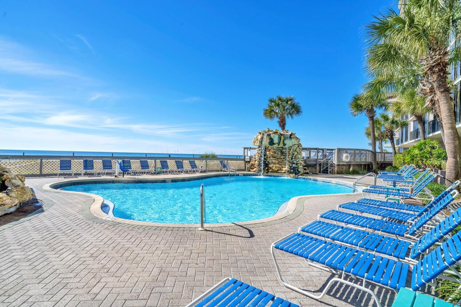 A swimming pool with blue lounge chairs, a stone waterfall feature, and a view of the ocean.