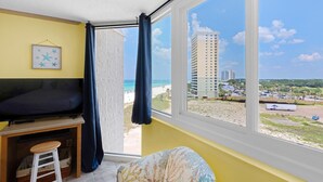 A room with a bed, a TV, a small stool, a window with a view of the beach and buildings. - Top of the Gulf 822 (Panama City Beach)