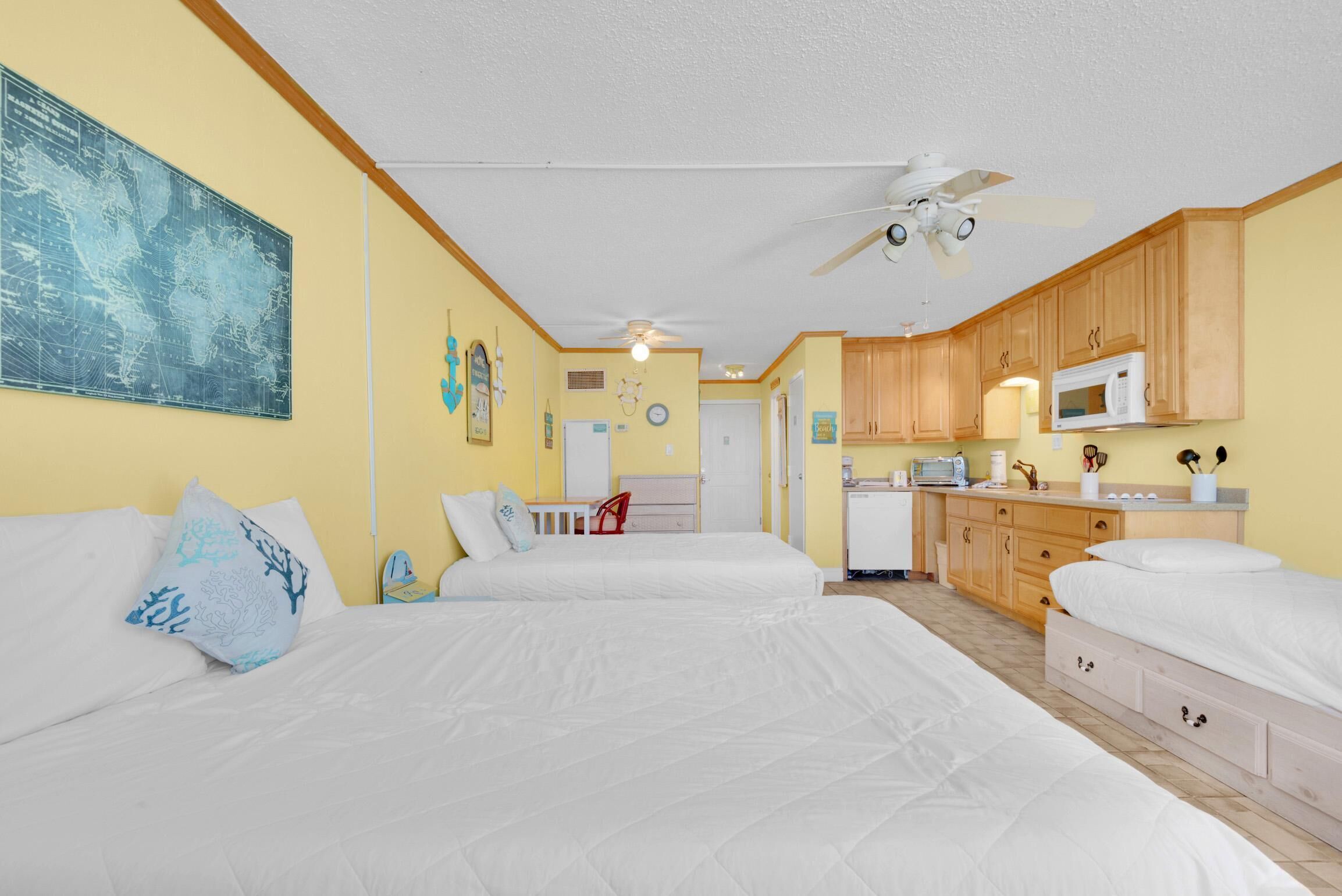 A hotel room with two beds, a kitchenette, a ceiling fan, and a world map on the wall.