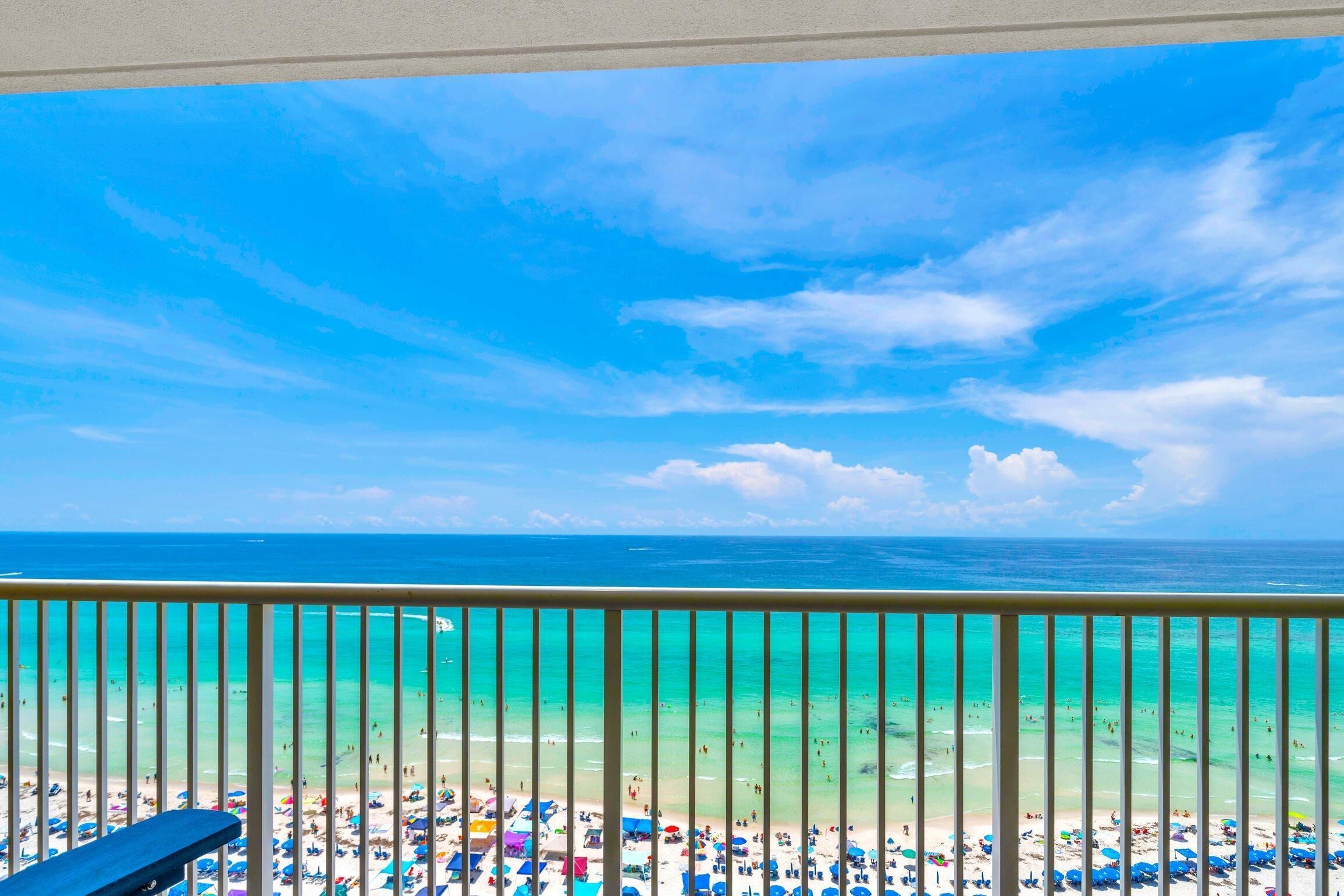 A beach view from a balcony with a railing, clear turquoise water, and a partly cloudy sky.