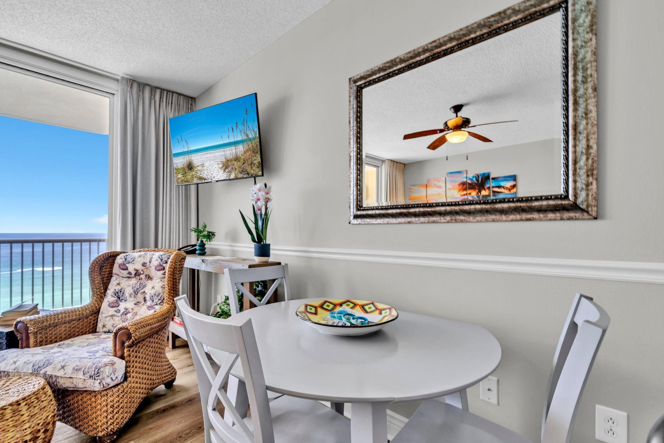 A balcony with a view of the ocean, a wicker chair, a white table with a decorative plate, and a ceiling fan.