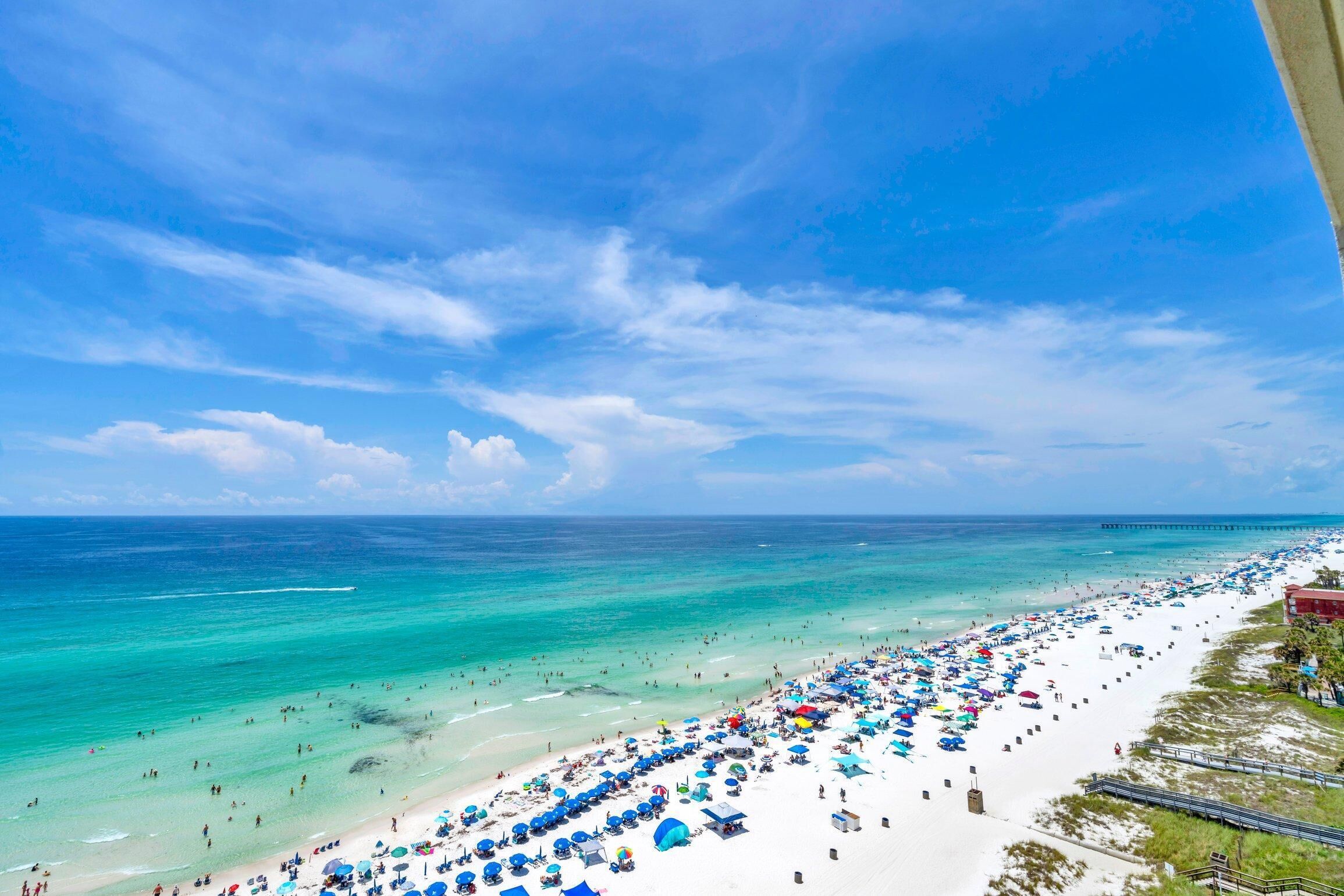 A beach with numerous umbrellas and people, a pier in the distance, and a clear blue sky.