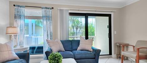 Sala de estar con un sofá azul, una mesa de centro blanca y una planta. Hay una puerta corrediza de vidrio que da a un balcón.