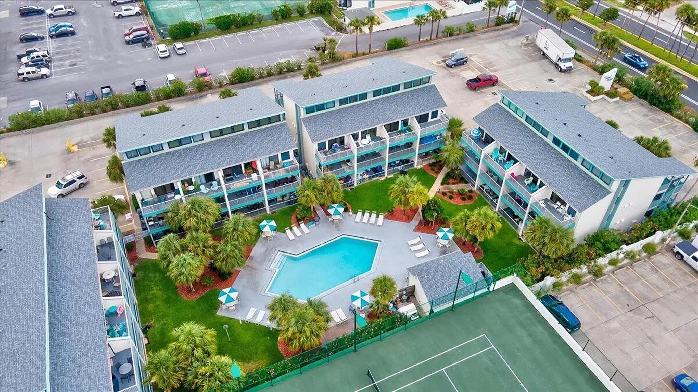 A multi-story hotel with a central courtyard featuring a swimming pool, sun loungers, and a tennis court.
