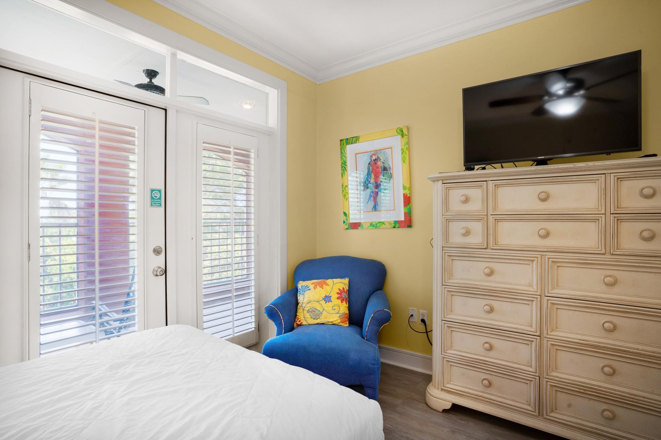 Une chambre à coucher avec un lit, un fauteuil bleu, une commode et un téléviseur fixé au mur.