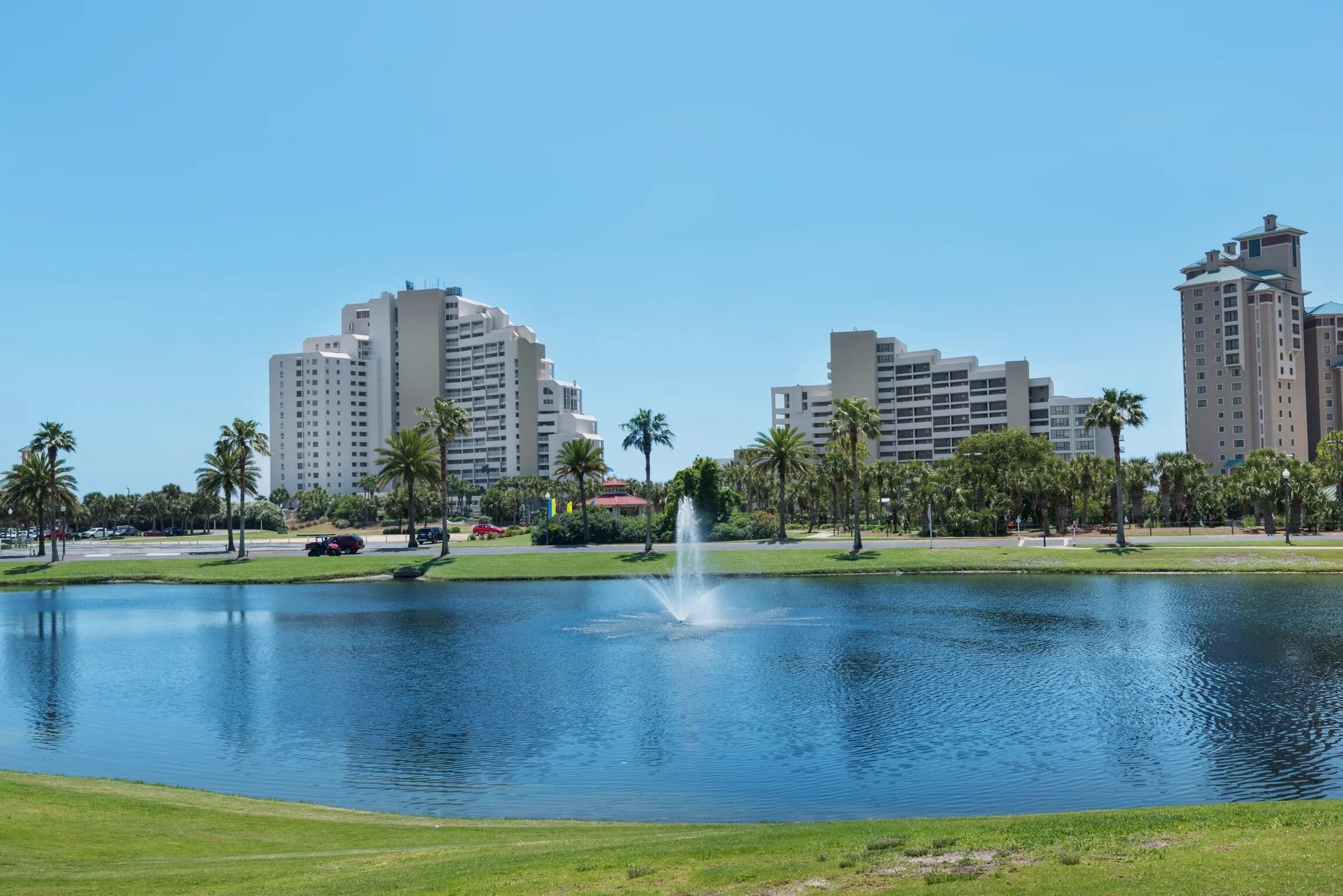A large body of water with a fountain in the center, surrounded by green grass and palm trees. In the background, there are tall buildings with multiple floors.